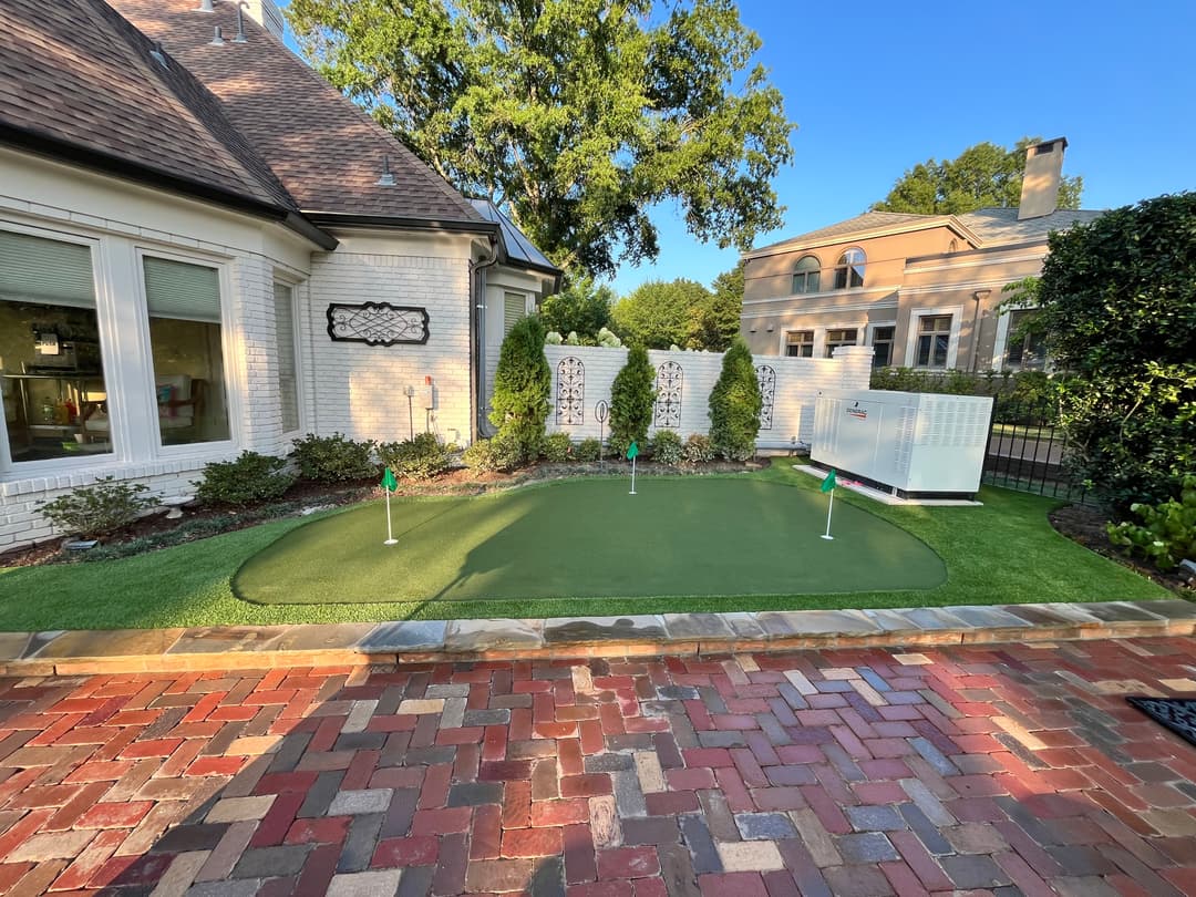 Backyard artificial turf putting green installation in Memphis, TN bordered by brick paver patio, creating a clean, low-maintenance golf practice area with proper drainage and finished hardscape edges. surrounded by landscaping and a brick patio.